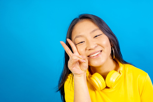 Studio photo with blue background of a smiling chinese woman with headphones gesturing peace sign