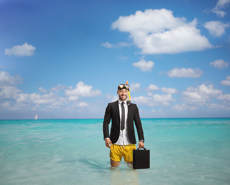 Businessman in a swimsuit with snorkelling fins and mask holding a briefcase and standing in the sea