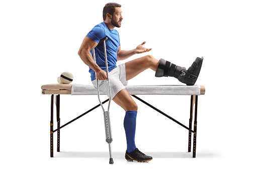 Football player with a crutch and injured foot sitting on a physical therapy bed isolated on white background