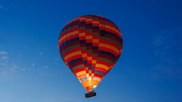 Photo of A colorful balloon in the clear dark sky at dawn, with fire burning inside, and people in the basket expressing joy.