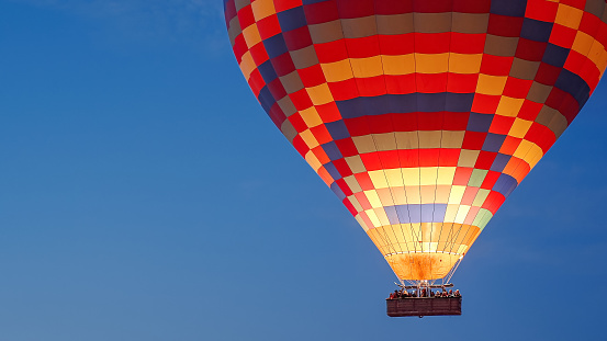 A multicolored hot air balloon with people in the basket, against the dark sky at dawn. Fire burns inside.