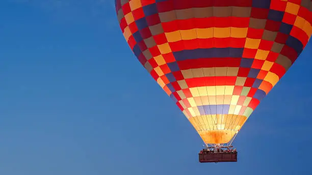 Photo of A multicolored hot air balloon with people in the basket, against the dark sky at dawn. Fire burns inside.