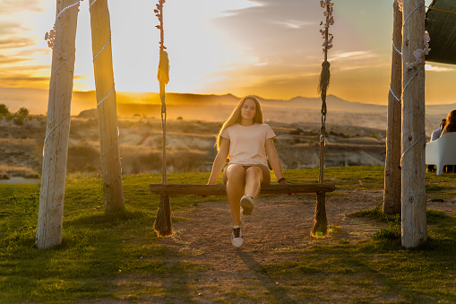 Young woman on a swing in the mountains against the backdrop of the sunset. The sky is of stunning colors, she is traveling and happy.