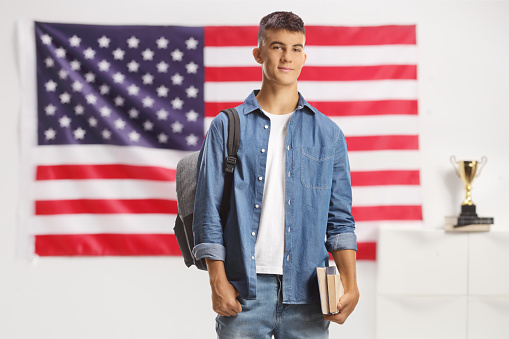 Male student with a backpack standing and holding books in front of american flag