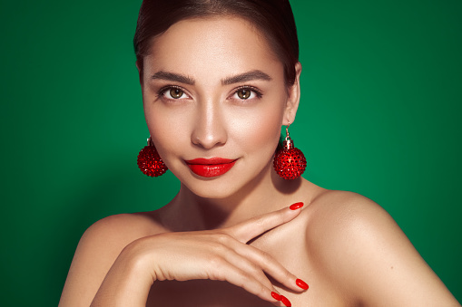 Portrait of beautiful young woman on green background, red lipstick and festive makeup, Christmas toy earrings, shiny glowing skin, Winter holidays concept
