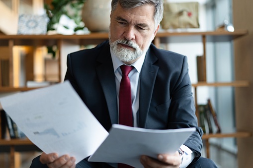 Mature businessman in formal suit concentrating on reading the report, sitting in the lobby and preparing for the meeting