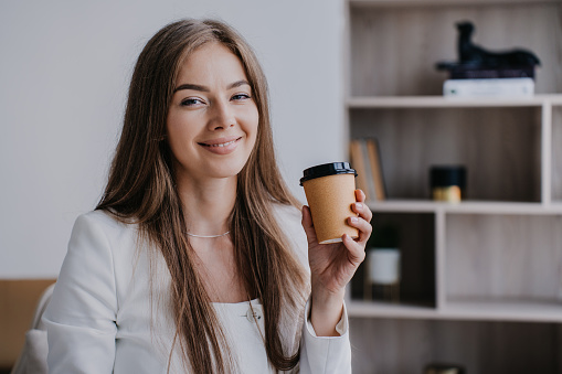 Beautiful American young businesswoman sitting at office enjoying coffee at morning. Long haired caucasian woman looking at camera happily smiling against bookshelves at home. Financial business.