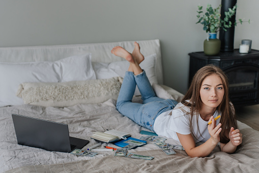 Purposeful female freelancer laying on bed with laptop diary dollar banknotes looks at camera. Pretty caucasian video blogger planning new podcast. Entrepreneur works at home.