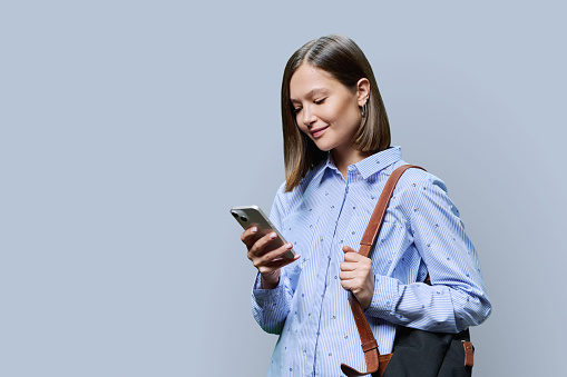 Young woman student with smartphone backpack on grey studio background. Smiling attractive female looking at phone. Using mobile applications apps for work business study leisure, technology, people