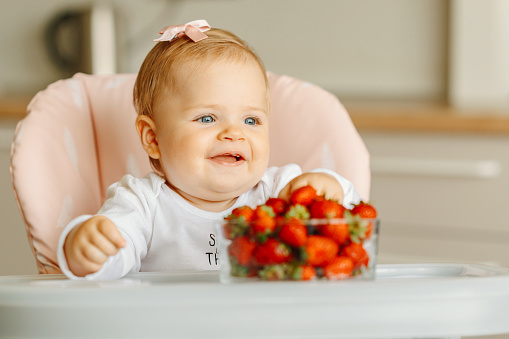 Photo of charming and happy baby girl eating strawberries in kitchen.
