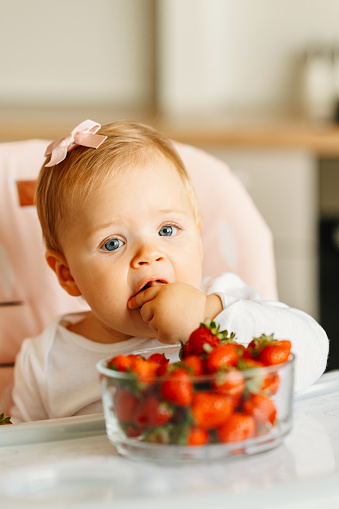Photo of charming and happy baby girl eating strawberries in kitchen.