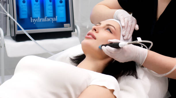 istock Cosmetology clinic. Professional female cosmetologist doing Hydradermabrasian procedure while being a work. Attractive nice woman lying on the medical bed while having beauty procedures 1820029585