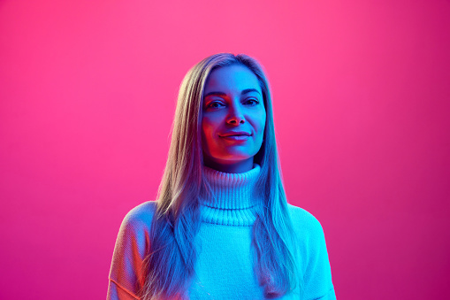 Close up portrait of young attractive calm woman in knitted warm sweater against pink background in neon light. Concept of beauty, human emotions, facial expression, fashion and style.