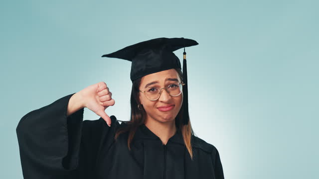 Thumbs down, graduation and student loans with a woman in studio on a blue background for education. Portrait, scholarship and debt with an unhappy university or college graduate looking upset
