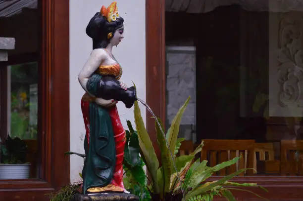 Side View Of A Statue Of A Woman Holding A Jug With Water Flowing In The Garden Around The Restaurant