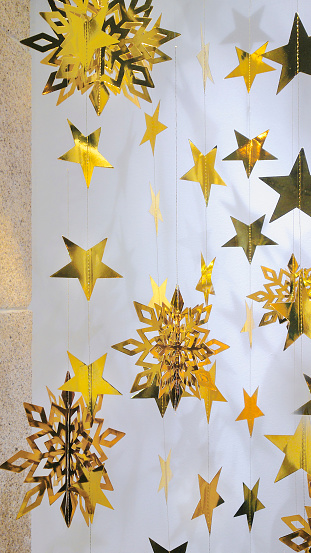 Gold colored star and snowflake shape figurines hanging, white wall background. Christmas decoration. Galicia, Spain.