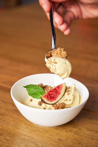 Vertical photo of a spoon grabbing some prickly pear mousse with gofio crumble served with mint and fig to decorate in a bowl in a wooden surface