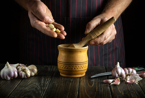 Chef adds the garlic to a wooden mortar before crushing. Close-up of a cook hands while working. Peasant food for national dish.