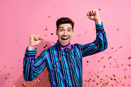 Portrait of overjoyed attractive guy raise fists success achievement flying confetti isolated on pink color background.