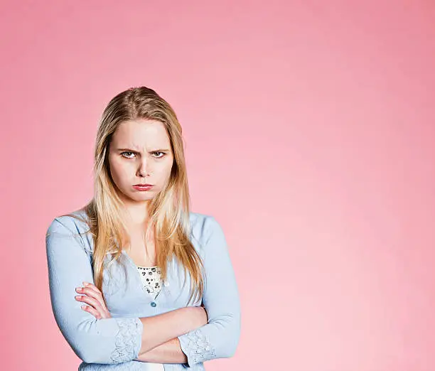 "This beautiful young blonde woman folds her arms and pouts, frowning and sulky. Rose pink background."