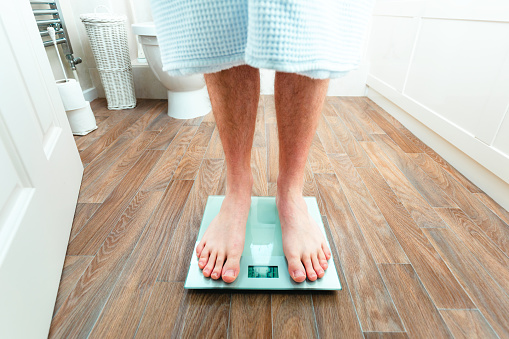 Color image depicting a man (specifically his legs and feet) weighing himself on the electronic weight scale at home.