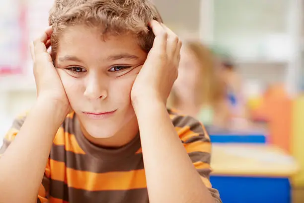 "Young boy at his desk in class holding his head, looking sad and lonely - copyspace"