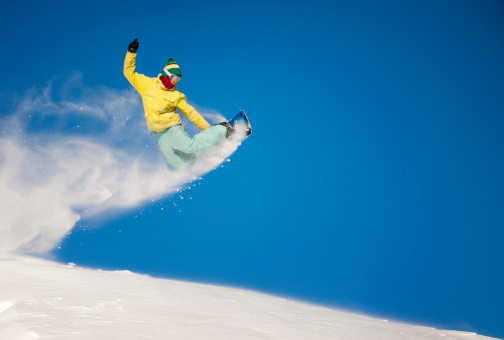 A professional snowboarder performing a backside air off the slopes against the deep blue sky with powder snow spraying.