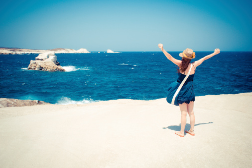 Young adult woman rejoices on a white rock looking the sea. The photo is taken near the fantastic beach of Sarakiniko in Milos, Cyclades Islands, Greece. Concept of freedom. Elaborated image for the vintage mood.