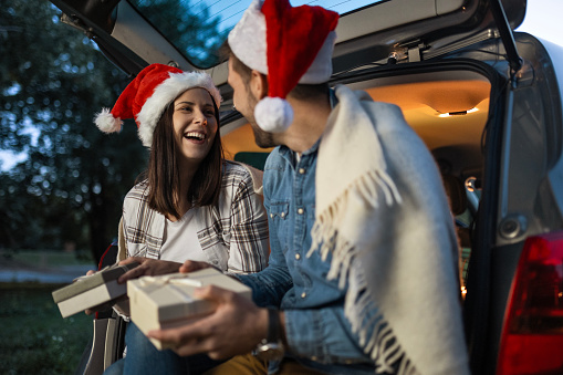 Happy couple is wearing Santa Claus hat and sitting in car trunk while enjoying moments of Christmas