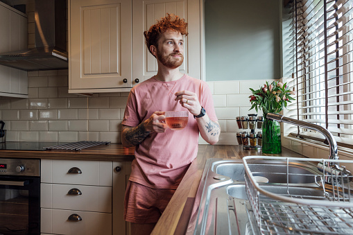 Three quarter length of a young redhead man standing in his kitchen on a morning, he is holding a cup of herbal tea and looking out the window at his home in County Durham, England. He's wearing a casual top and shorts.

Video also available for this scenario.