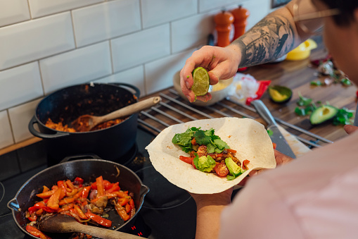 Over the shoulder view of a young unrecognisable LGBTQI+ male couple standing in their kitchen, they have prepared chicken and vegetables at their home in County Durham, England. One man is squeezing lime juice onto the vegetables and chicken in a soft taco.\n\nVideo also available for this scenario.