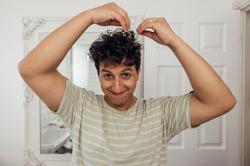 Front view close up of a young man with curly hair standing in his bathroom on a morning, he is adding pomade to his hair while styling it and looking at the camera at his home in County Durham, England. He's wearing a casual top.

Video also available for this scenario.