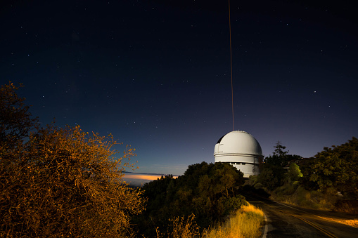 The mount hamilton observatory at night with stars in the night sky. A laser is shooting up out of the observatory dome.