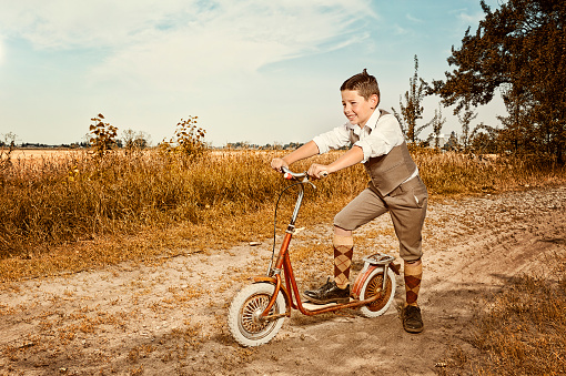 Little boy riding his scooter down a country lane, retro-style.  Happy and smiling.  Looking off camera and forward to him.  Cowlick sticking straight up on the top of his head just like the movie character.  Country, urban scene.  Great for back to school ads for children.