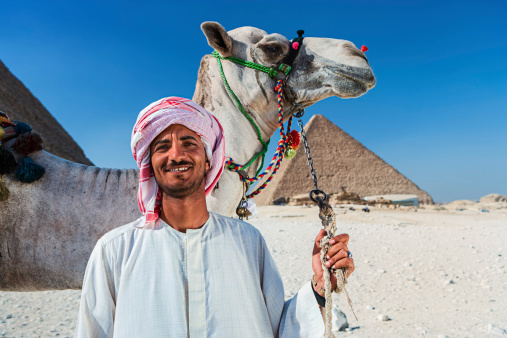 Giza, Cairo, Egypt - September 30, 2021: Egyptian in national dress sits astride a camel and smiles. Tourists walk and take pictures near the Egyptian pyramid.