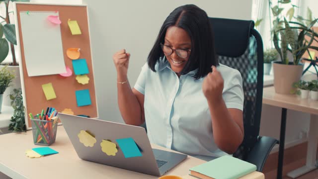 Beautiful smart black businesswoman sitting at home office desk, raising arms