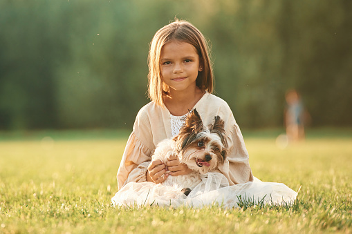 Front view, sitting together. Cute little girl is on the field with dog.