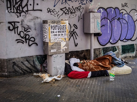 Homeless young man sitting on sidewalk and begging alone. He is wearing protective face mask and holding out a can and begging for money. His head is bowed in shame and tiredness and hunger.