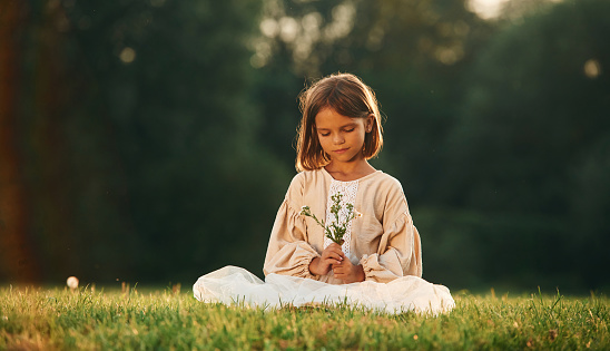 Sitting, holding little flowers. Little girl is on the summer field outdoors.