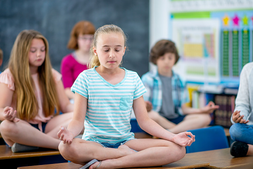 A small class of elementary students sit on to p of their desks with their legs crossed as they meditate.  They are each dressed casually as they hold a lotus pose and focus on their breathing.