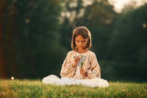 Sitting, holding little flowers. Little girl is on the summer field outdoors.