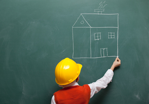 Back view photo of little boy wearing  a yellow helmet drawing a house on blackboard.He is wearing an orange waist and a white shirt.His face is turned to blackboard.He is drawing with right hand via chalk.He is located on the down centre of frame.The photo was shot with a full frame DSLR camera. 