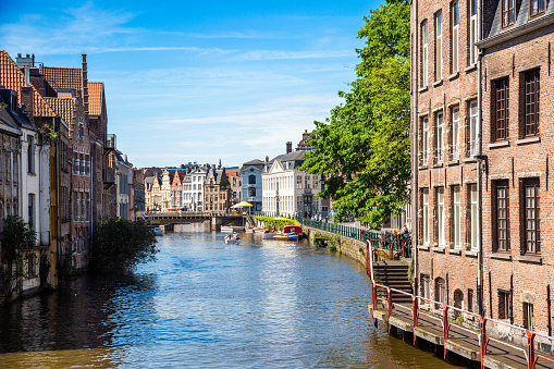 A canal in Ghent, Belgium with historic brick buildings on the left and a blue sky in the background. The buildings have orange and yellow facades and are in various architectural styles. The canal is lined with trees and there are boats on the water. Bridge and more buildings is in a distance.