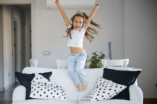 Carefree little girl dancing on the sofa bed. She is listening music via wireless headphones.