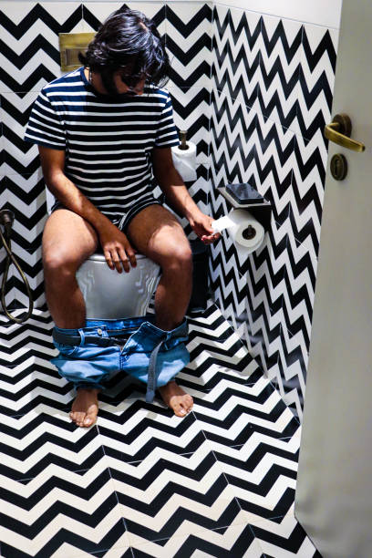 Image of Indian man sitting on toilet pulling paper off toilet roll holder, denim jeans around ankles, luxury hotel bathroom with mobile phone charging base, black and white zig-zag patterned walls and floor tiles Stock photo showing a hotel bathroom featuring a white toilet with modern fittings include bathroom hygiene in the form of a hand shower bidet head attachment, which has been fixed to the wall next to the WC, complete with wall bracket and brass hose connection. pedal bin stock pictures, royalty-free photos & images