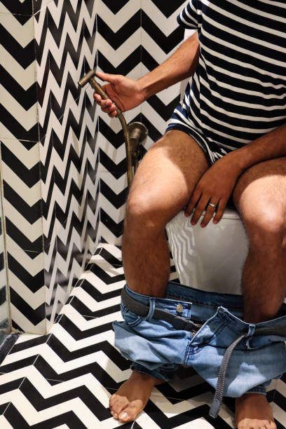 Close-up image of unrecognisable man sitting on toilet holding brass hand shower bidet head attachment, denim jeans around ankles, luxury hotel bathroom with black pedal bin, black and white zig-zag patterned walls and floor tiles Stock photo showing a hotel bathroom featuring a white toilet with modern fittings include bathroom hygiene in the form of a hand shower bidet head attachment, which has been fixed to the wall next to the WC, complete with wall bracket and brass hose connection. pedal bin stock pictures, royalty-free photos & images