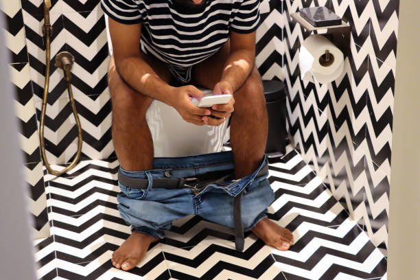 Close-up image of luxury hotel bathroom with unrecognisable man sitting on toilet looking at Smartphone, denim jeans around ankles, black pedal bin, black and white zig-zag patterned walls and floor tiles Stock photo showing a hotel bathroom featuring a white toilet with modern fittings include bathroom hygiene in the form of a hand shower bidet head attachment, which has been fixed to the wall next to the WC, complete with wall bracket and brass hose connection. pedal bin stock pictures, royalty-free photos & images