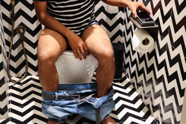 Stock photo showing a hotel bathroom featuring a white toilet with modern fittings include bathroom hygiene in the form of a hand shower bidet head attachment, which has been fixed to the wall next to the WC, complete with wall bracket and brass hose connection. imagen de primer plano del baño de un hotel de lujo con un hombre irreconocible sentado en el inodoro, jeans de mezclilla alrededor de los tobillos, base de carga de teléfono móvil, portarrollos de papel higiénico, paredes y baldosas estampadas en zig-