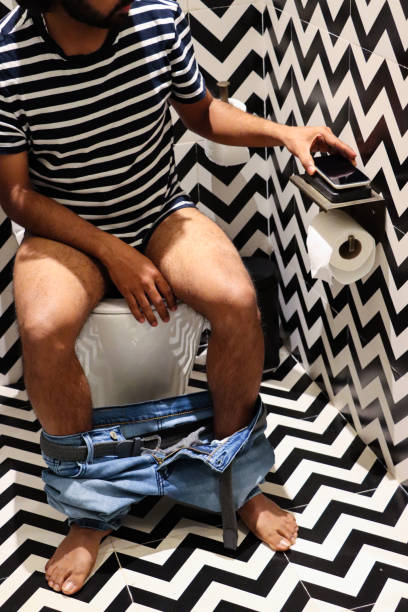 Image of luxury hotel bathroom with unrecognisable man sitting on toilet, denim jeans around ankles, mobile phone charging base, toilet roll holder,  black and white zig-zag patterned walls and floor tiles Stock photo showing a hotel bathroom featuring a white toilet with modern fittings include bathroom hygiene in the form of a hand shower bidet head attachment, which has been fixed to the wall next to the WC, complete with wall bracket and brass hose connection. pedal bin stock pictures, royalty-free photos & images