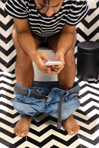 Close-up image of luxury hotel bathroom with Indian man sitting on toilet looking at Smartphone, denim jeans around ankles, black pedal bin, black and white zig-zag patterned walls and floor tiles Stock photo showing a hotel bathroom featuring a white toilet with modern fittings include bathroom hygiene in the form of a hand shower bidet head attachment, which has been fixed to the wall next to the WC, complete with wall bracket and brass hose connection. pedal bin stock pictures, royalty-free photos & images